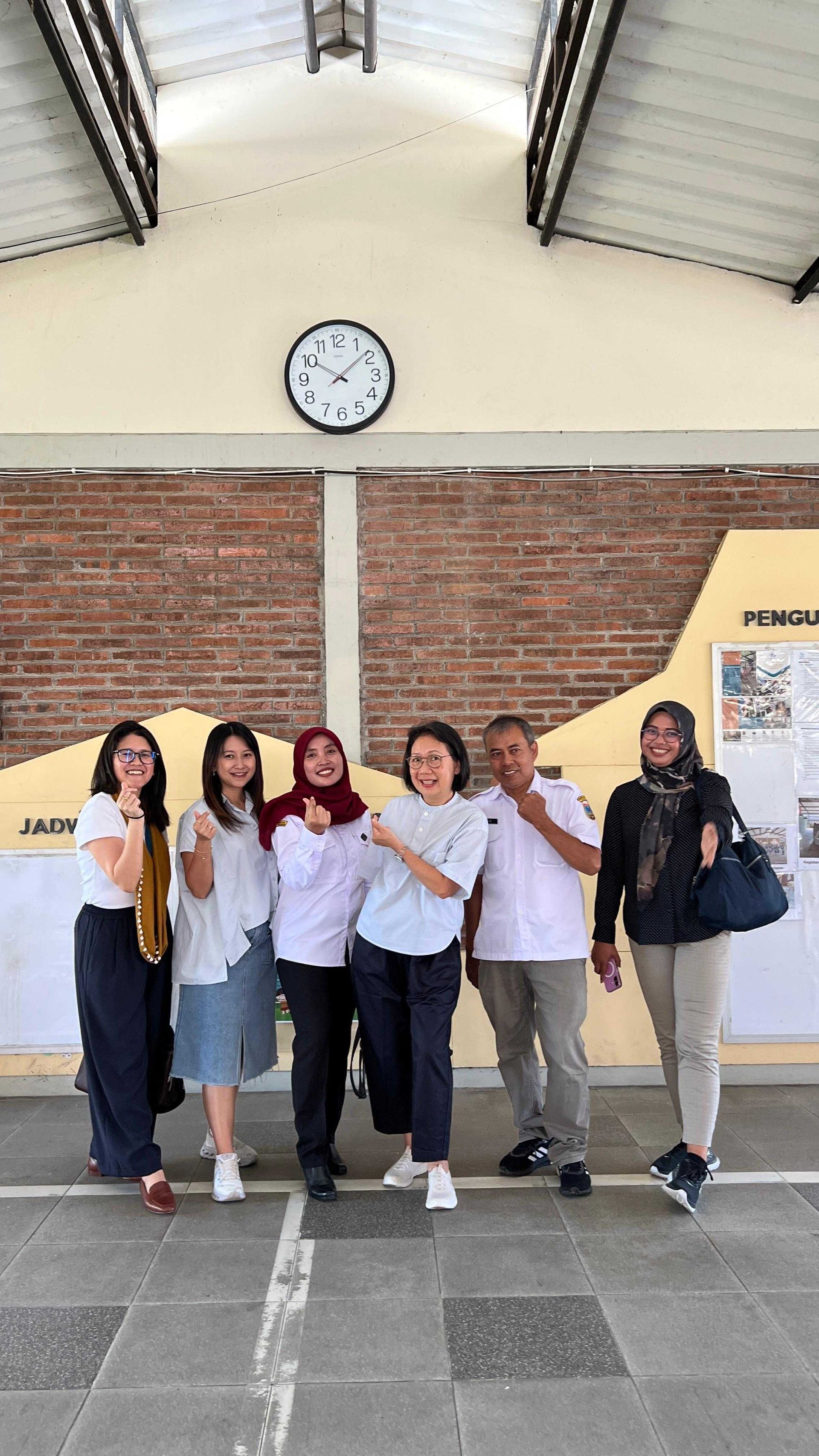 Sekelompok orang tersenyum, berpose di luar di bawah atap, dengan jam di dinding di belakang 