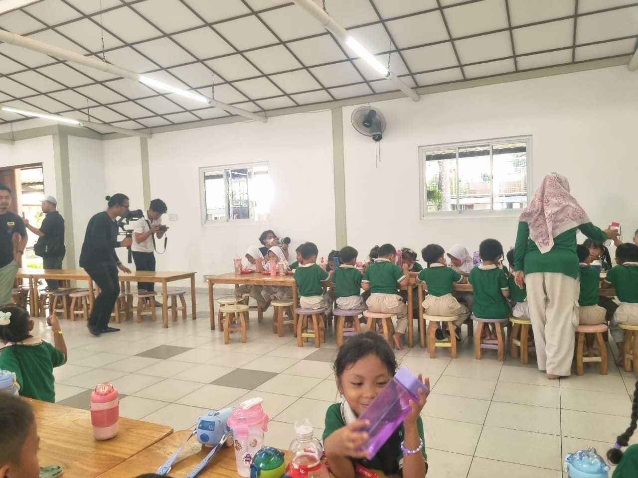 Children in green uniforms at tables in a brightly lit room, with adults, one taking a photo.