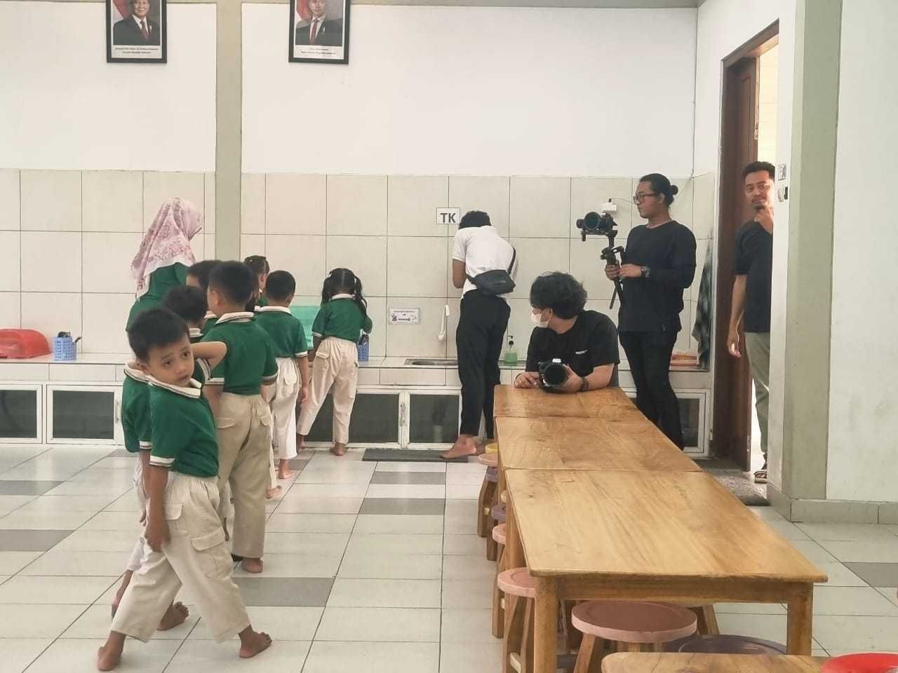 Children in green uniforms line up, facing cameras in a room with a long table and adults.