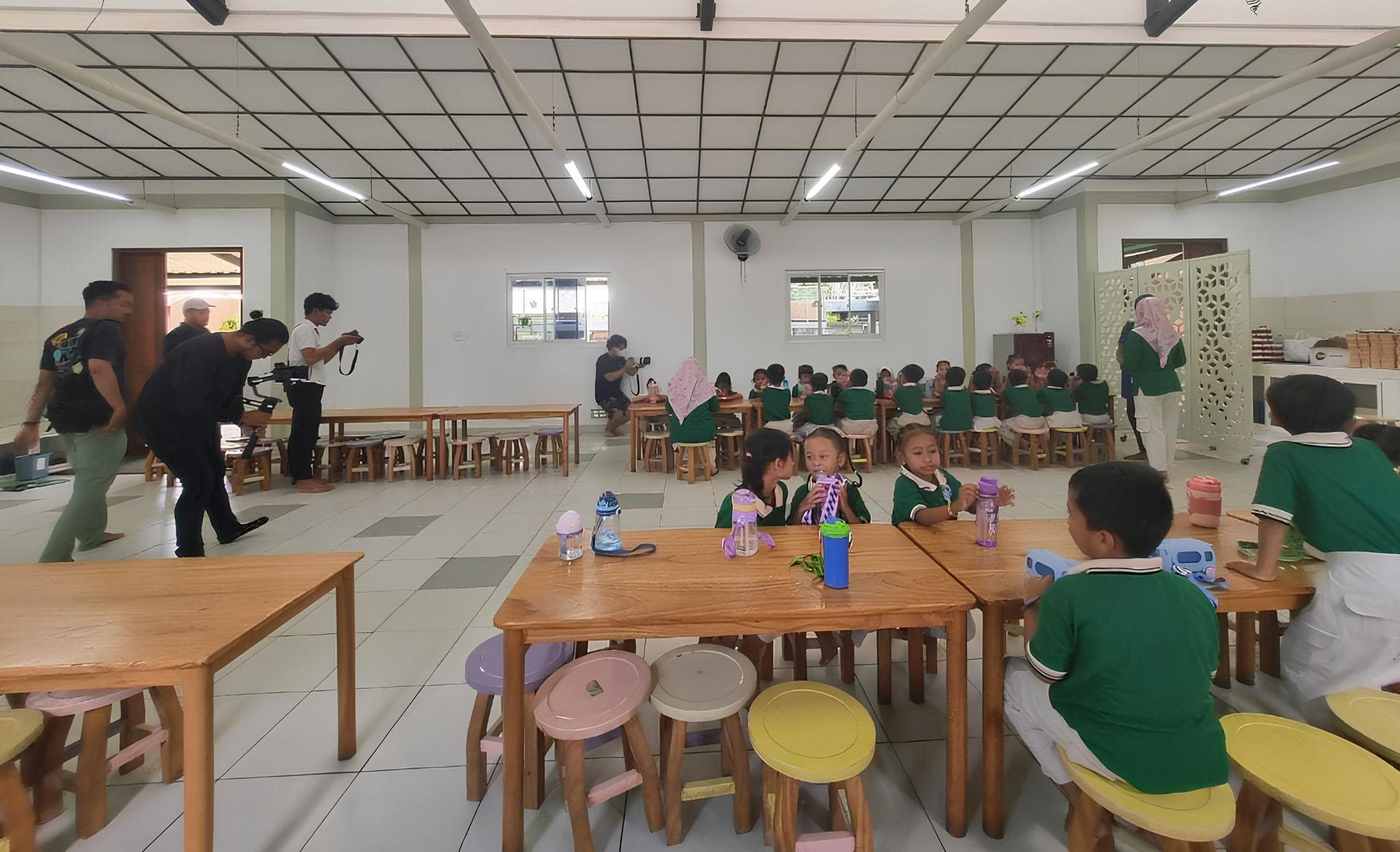 Children in green uniforms seated at small tables, some with adults, in a brightly lit room.