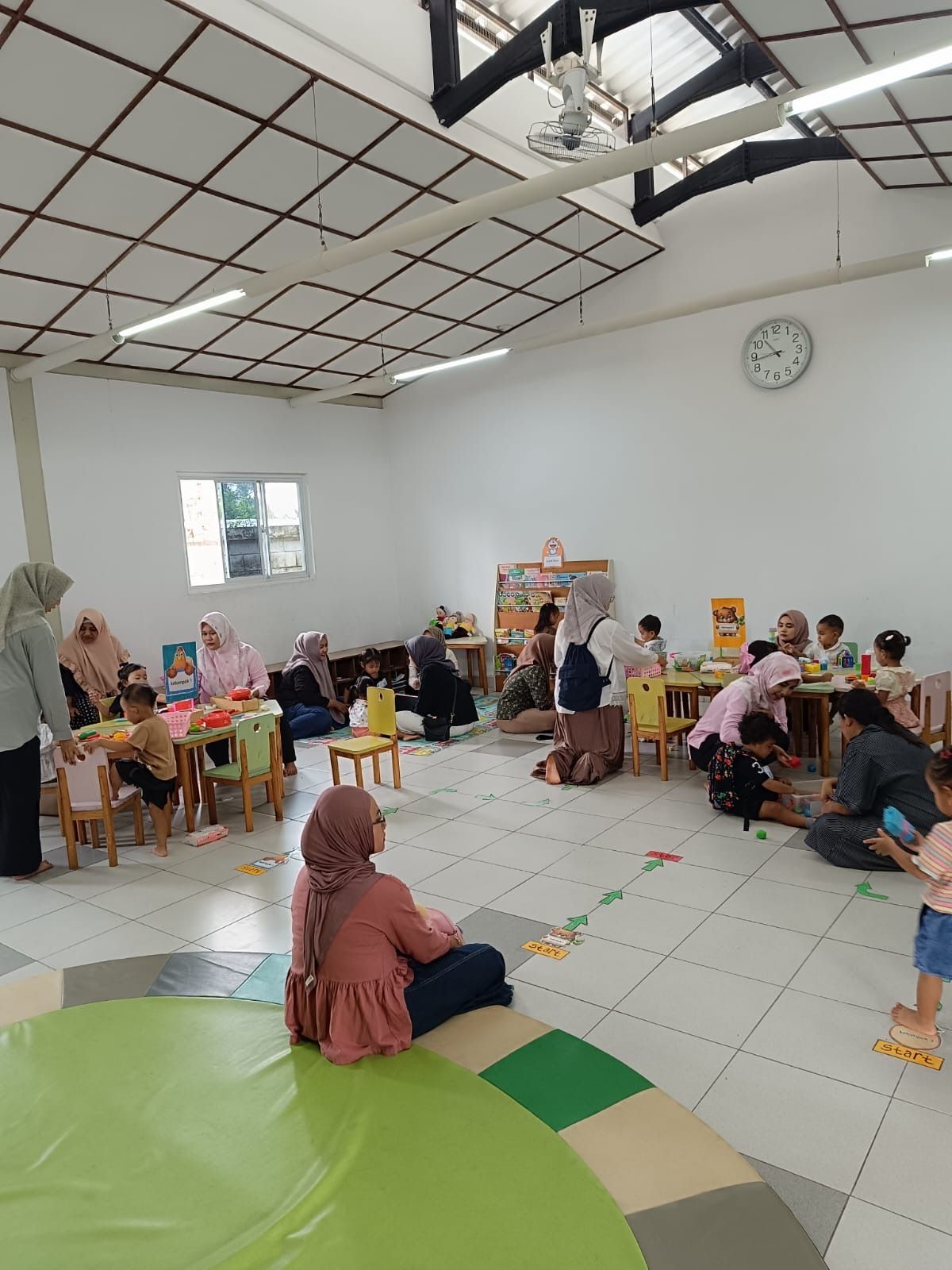 People gathered in a bright community room, seated on the floor and at small tables, with a green play mat in front.