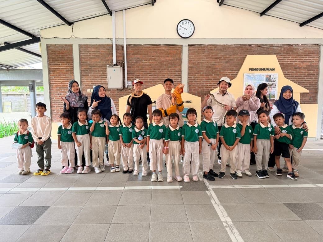 Students in green uniforms pose with teachers in a school hall, with a wall clock overhead.