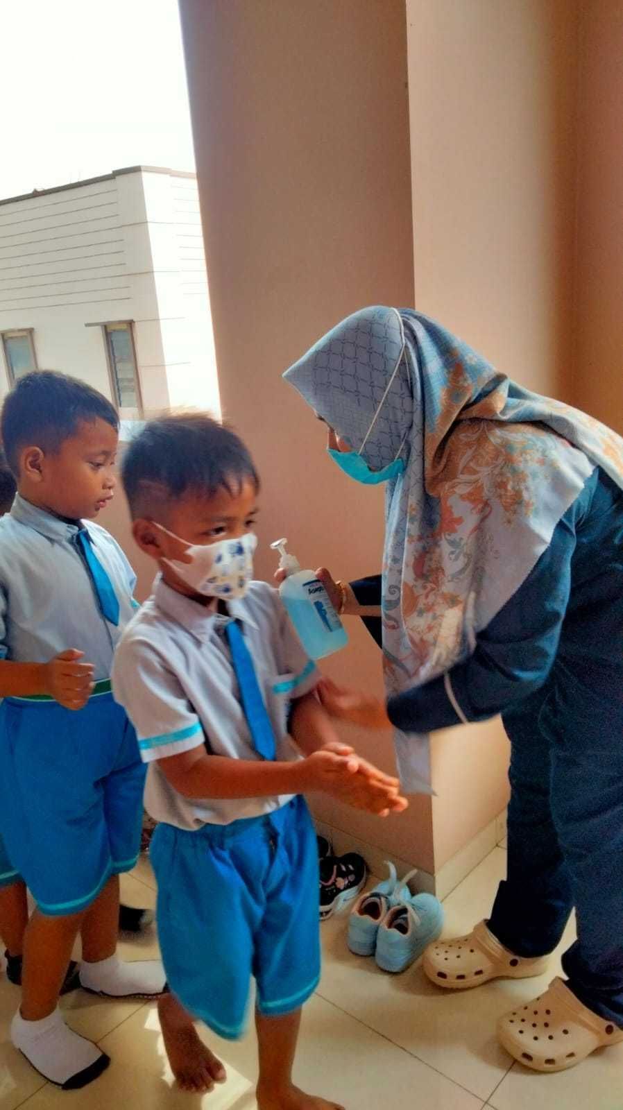 Teacher applying hand sanitizer to a child in a school uniform. Other children wait nearby. Outdoors.