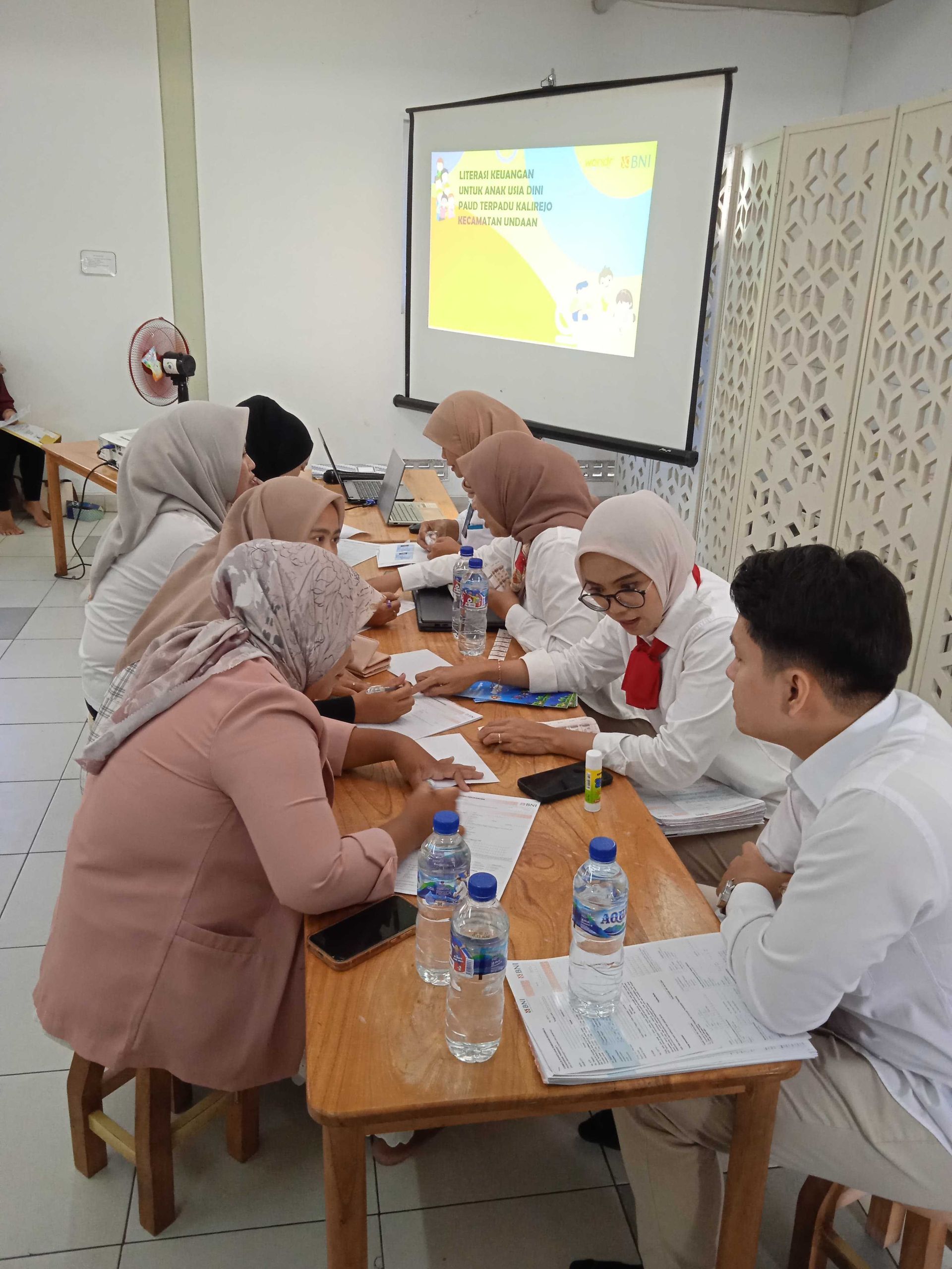 Group of people in headscarves and white shirts at a table, working on papers. Projector screen visible.