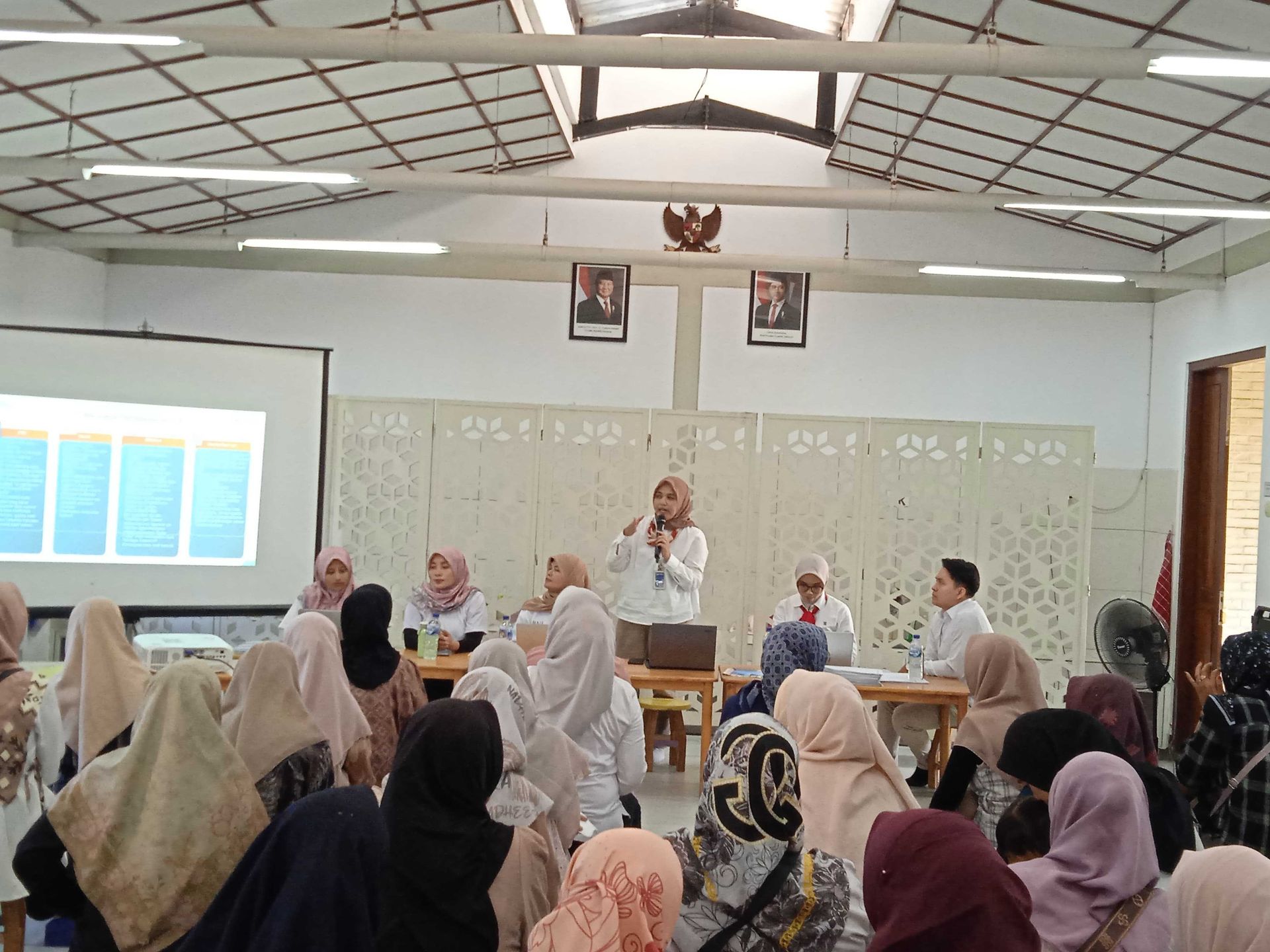 Sebuah presentasi sedang berlangsung. Seorang pembicara di podium di hadapan audiens perempuan