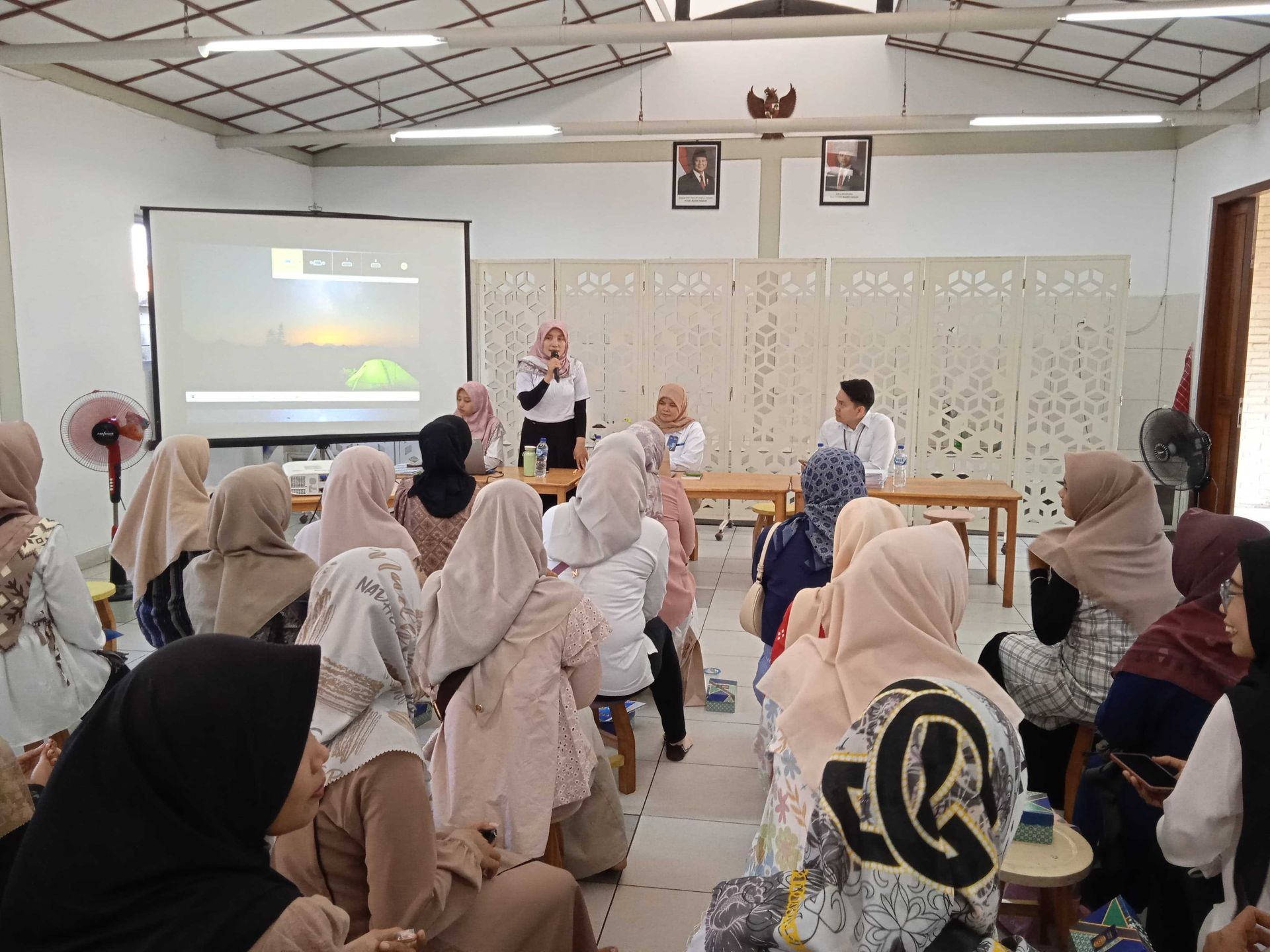 A presentation taking place in a room with women wearing headscarves; a presenter speaks at the front.