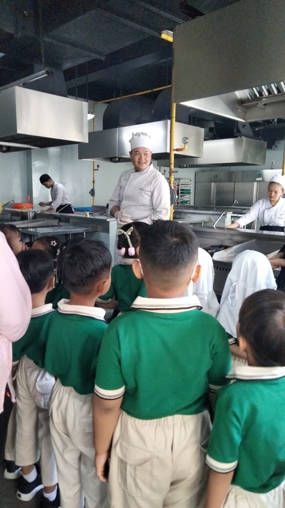 Children in green shirts watch a chef in a kitchen.
