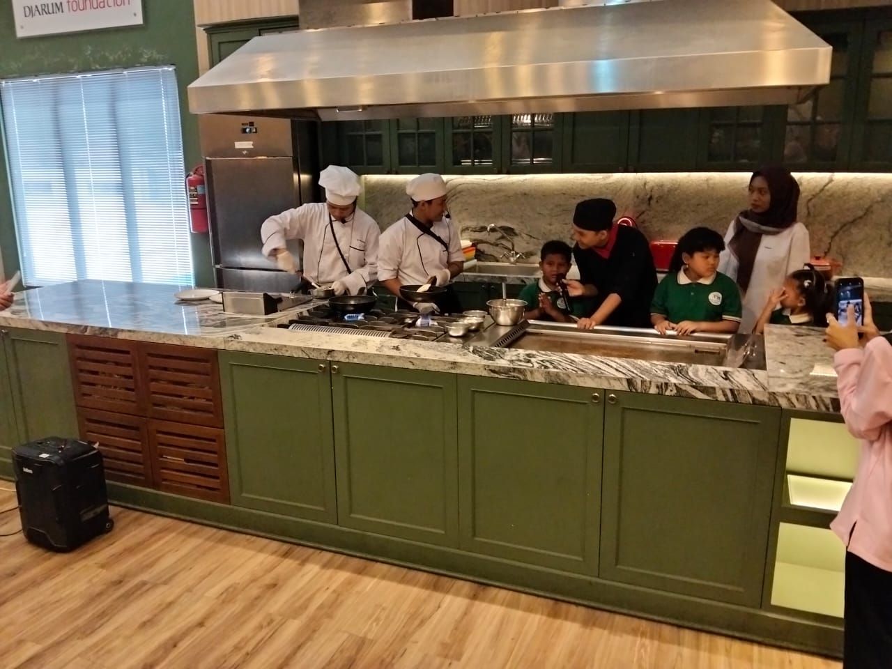 Chefs cooking at a counter in a green kitchen, with children and a woman watching.