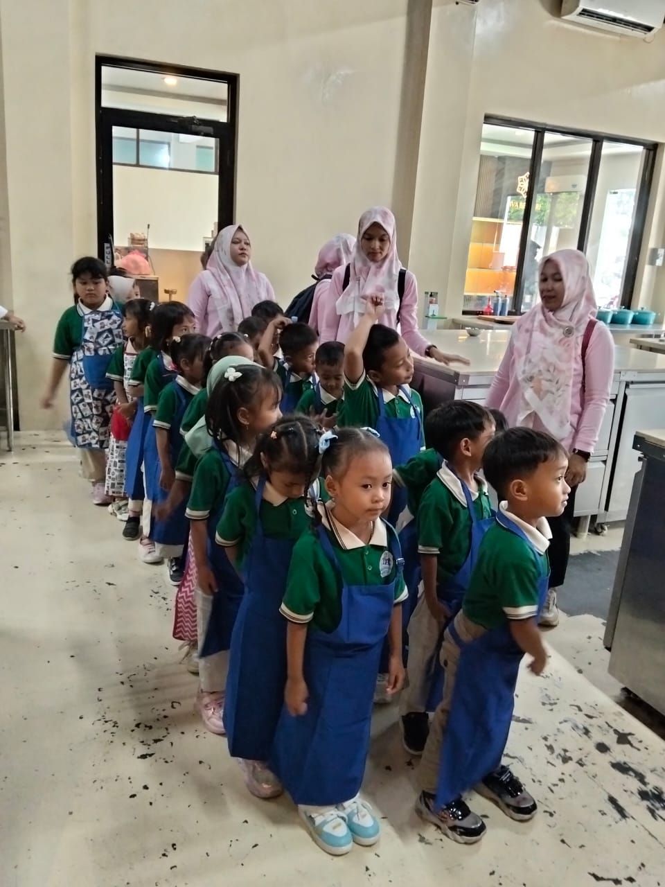 Children in green shirts and blue aprons line up in a kitchen, guided by teachers.
