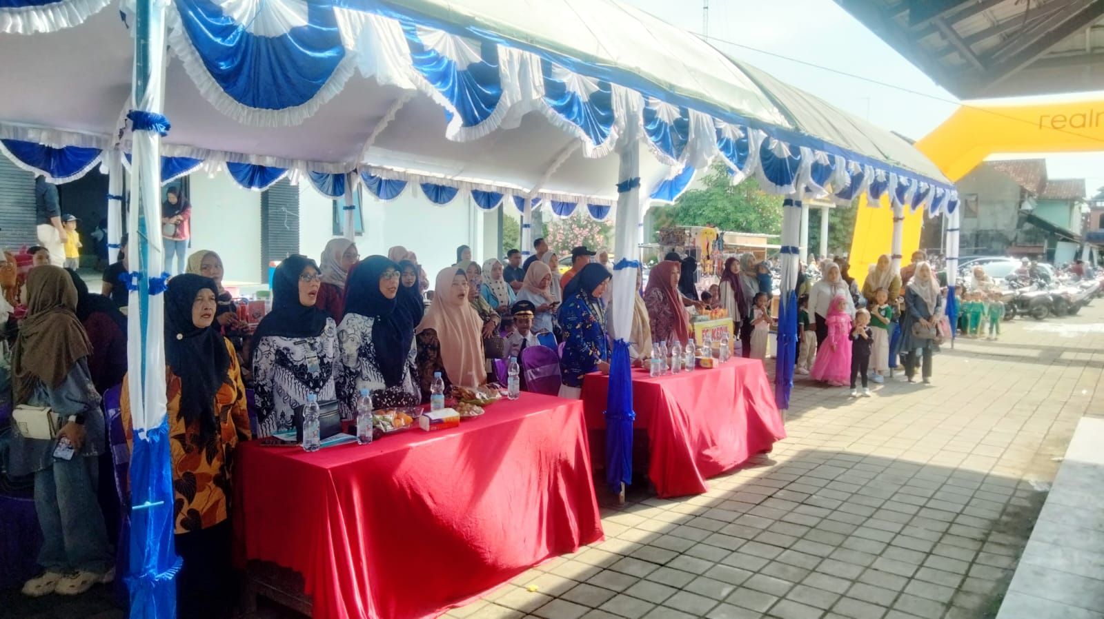 People standing behind tables covered in red cloth, under a blue and white tent.