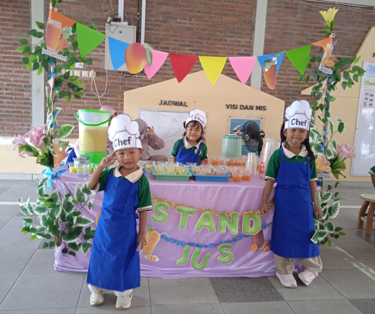 Three children in chef hats and aprons at a decorated stand selling drinks, smiling.