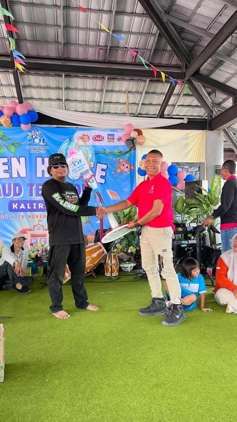 Two men exchanging items at an outdoor event. One hands the other a plate. Balloons and banners visible.