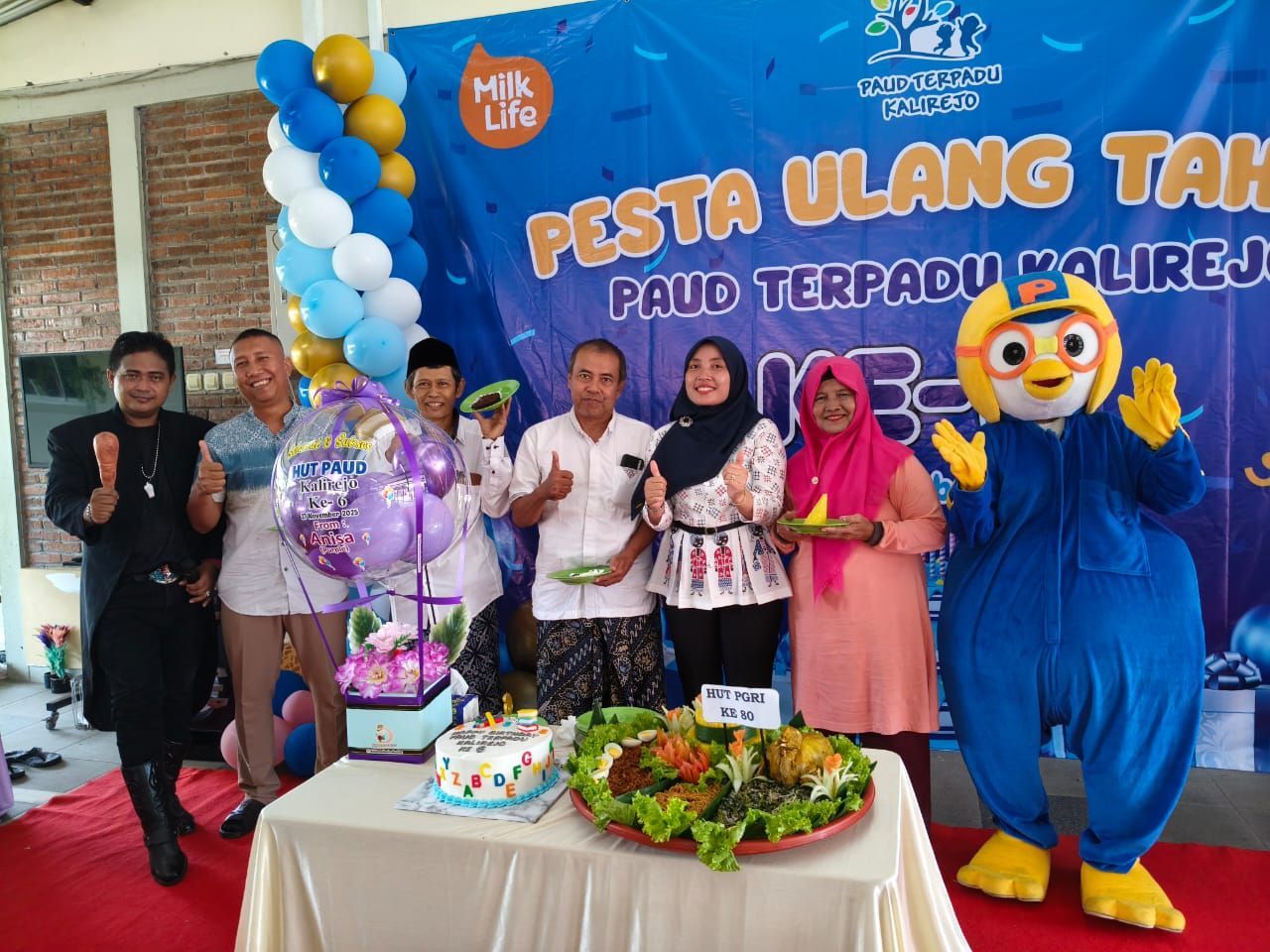 People celebrating a birthday with a banner, cake, balloons, and a mascot; they are outdoors.