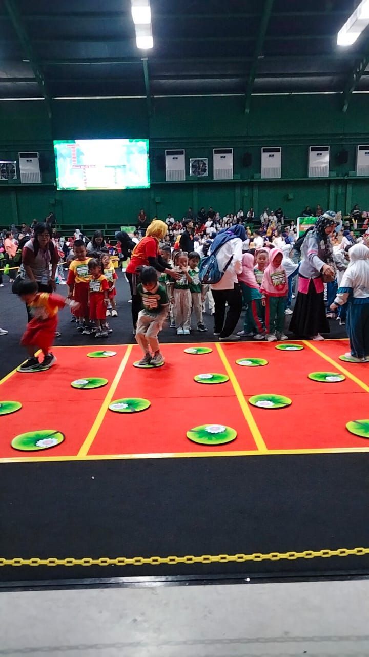 Children playing a game on a red mat with lily pad markers in an indoor event space, observed by adults.