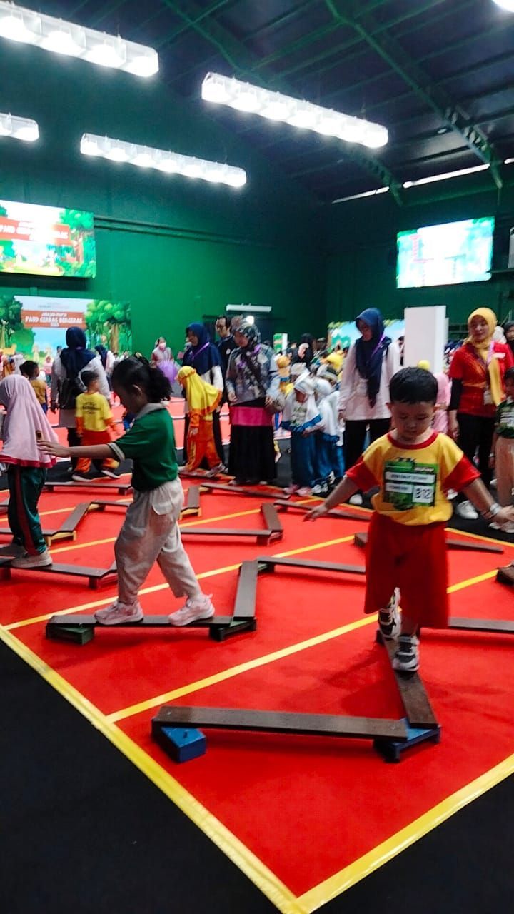 Children balancing on a wooden track in an indoor play area. Red carpet, other kids & adults watching.
