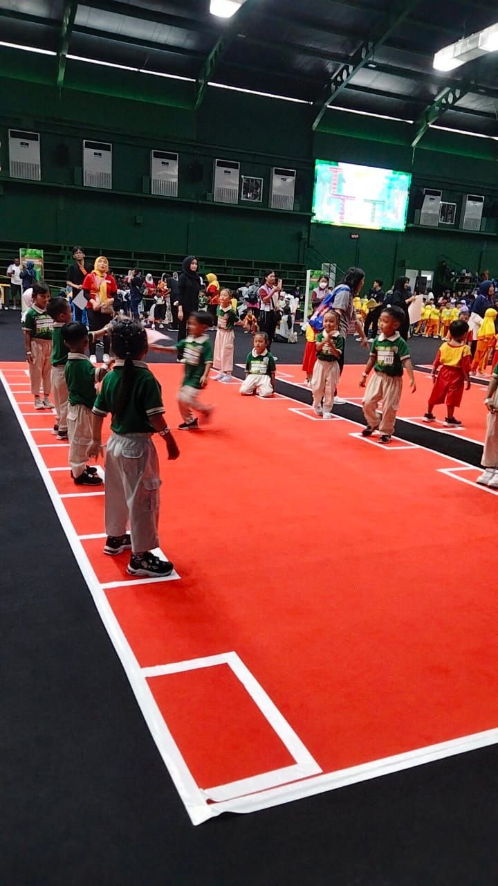 Children in green shirts and tan pants line up on red carpet. Indoor event.