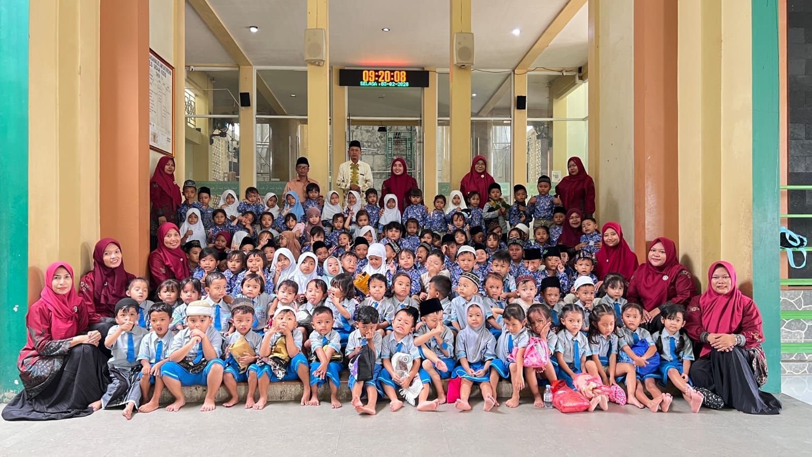 A large group of students and teachers in uniforms pose for a photo in front of a building with light-colored pillars.