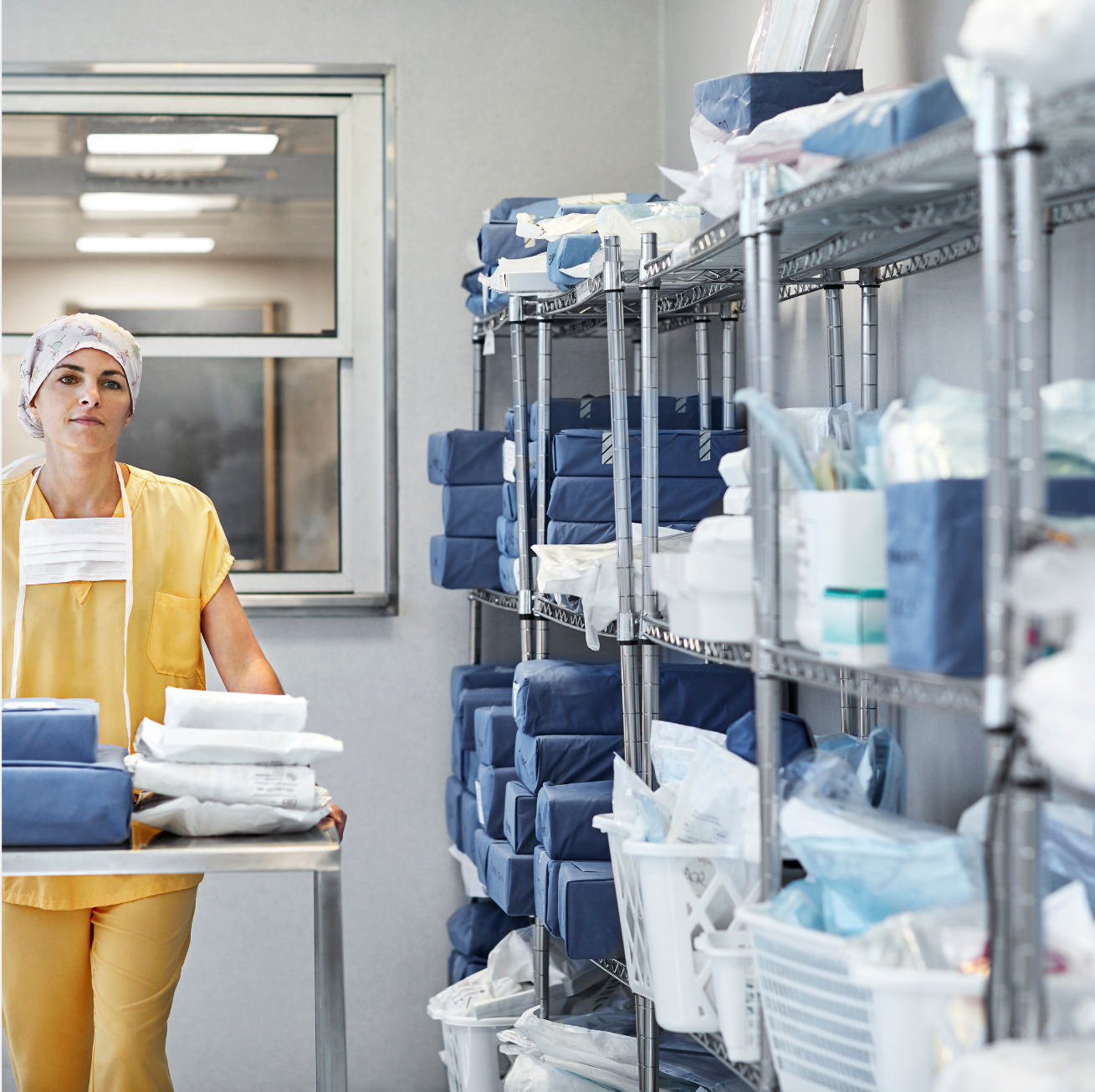 Woman in medical scrubs and cap, holding a tray of supplies, in a storage room with shelves of supplies.