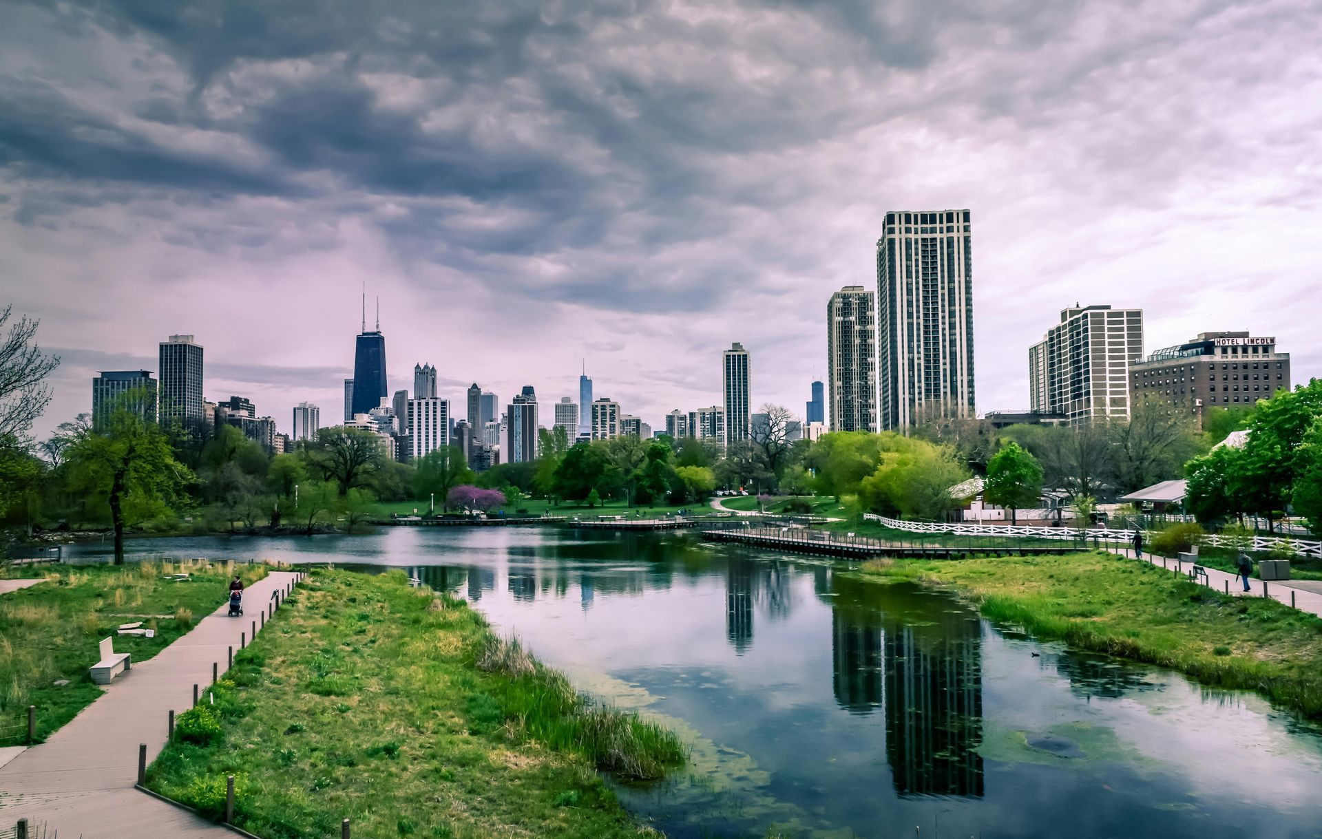 Chicago skyline view from a park with a reflective lake under a cloudy sky.