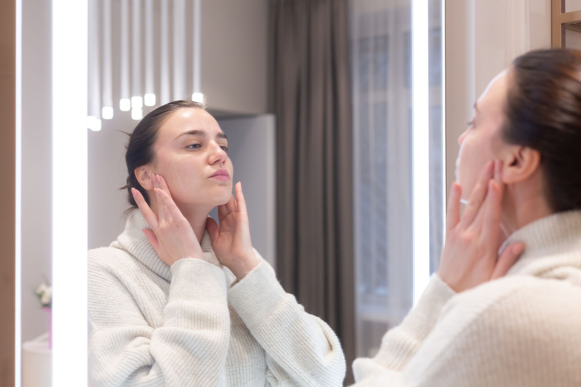 A woman is looking at her face in a bathroom mirror.