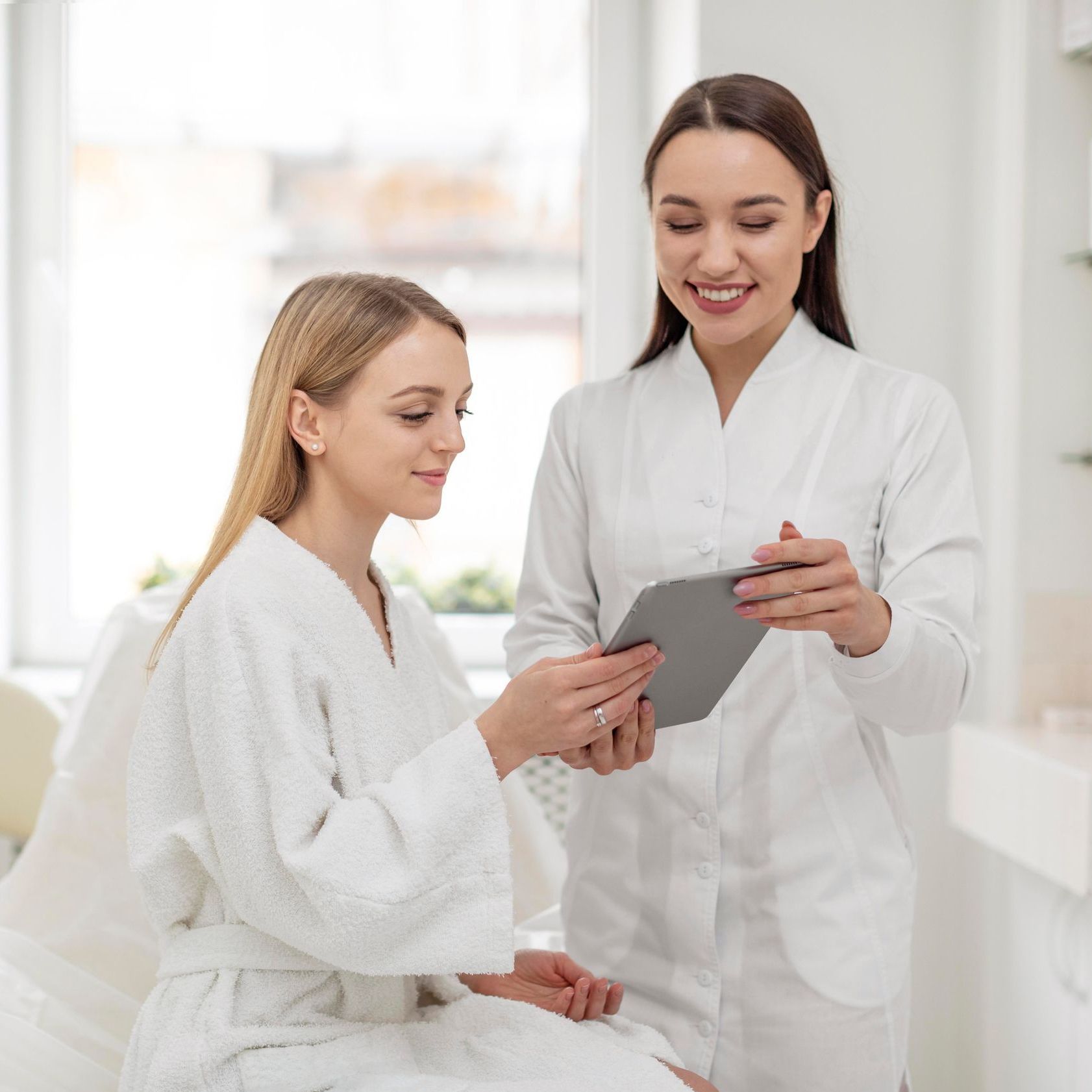 A woman in a bathrobe is sitting next to a woman holding a tablet.
