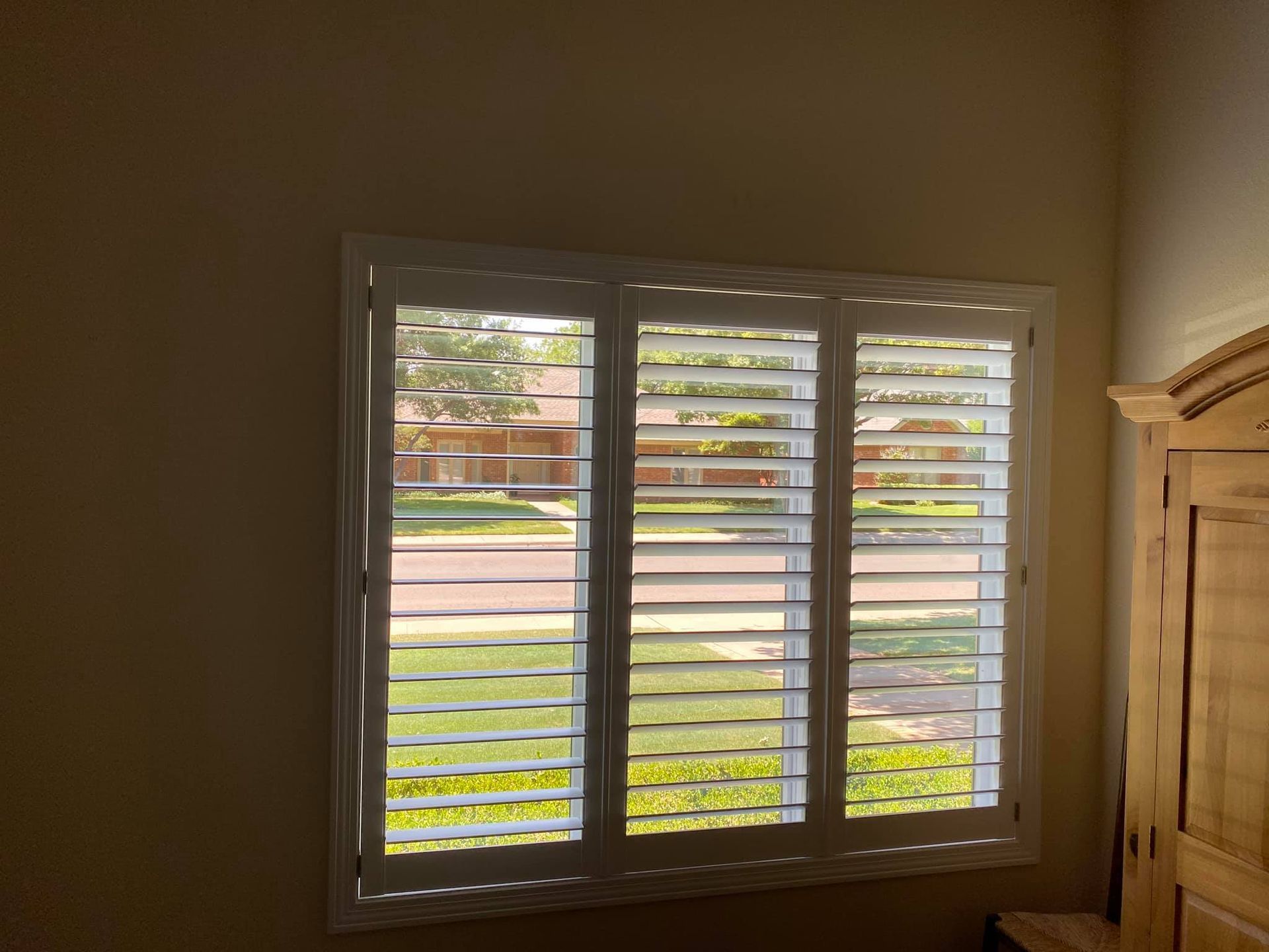A window with white plantation shutters partially open, showing a view of a street, lawn, and house across the way.