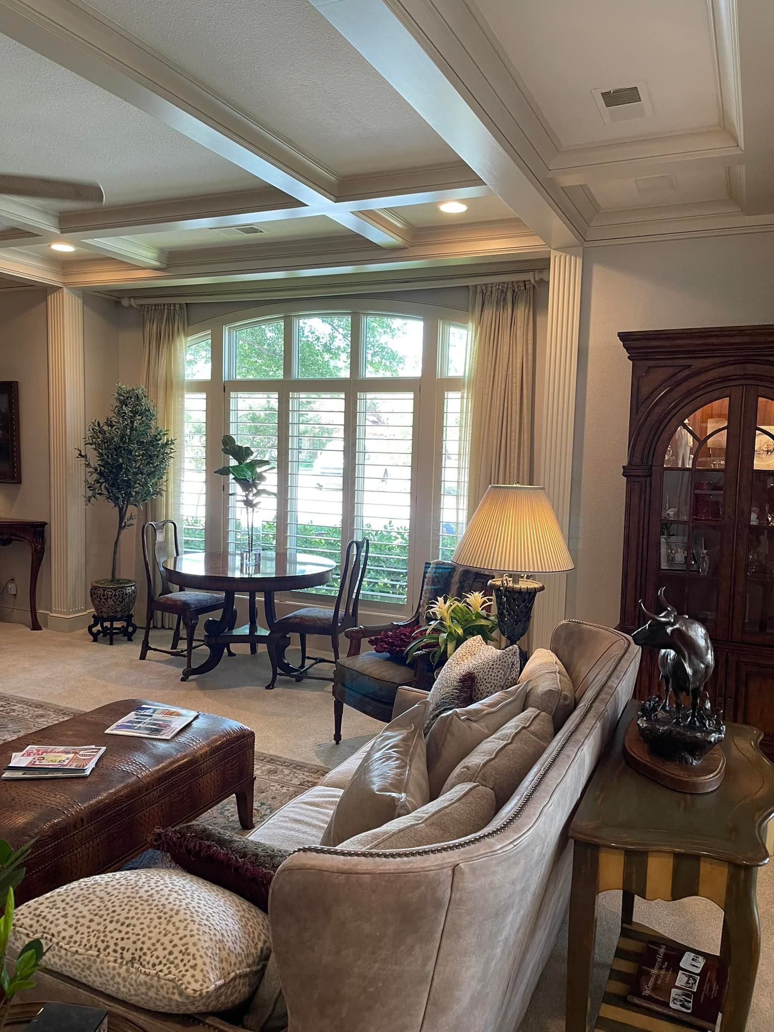 A formal living room featuring a tan sofa, a coffee table, a dining set by a large window, and a wooden hutch.