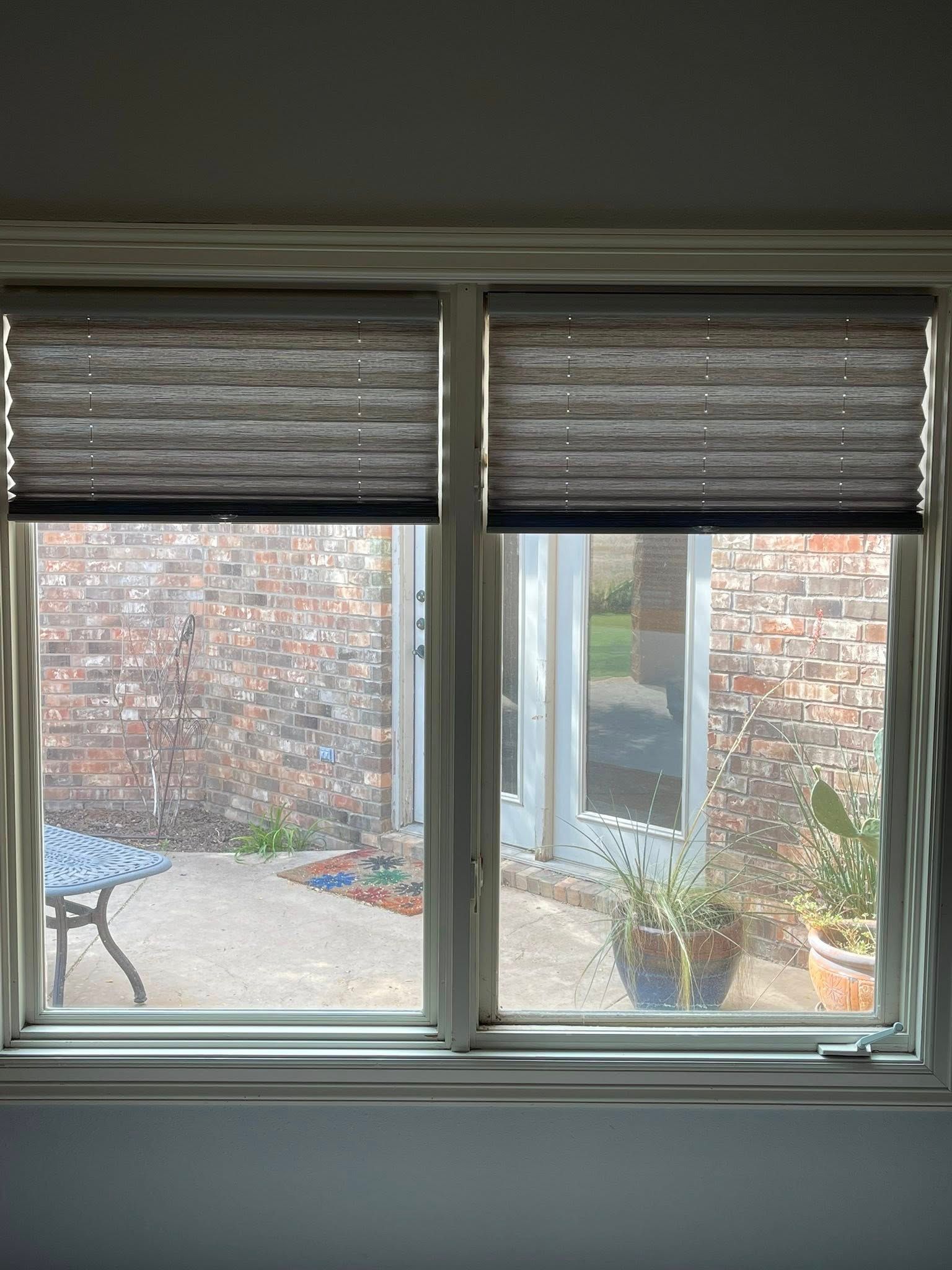A window with grey pleated shades partially lowered, overlooking a patio with a table, potted plants, and a brick wall.