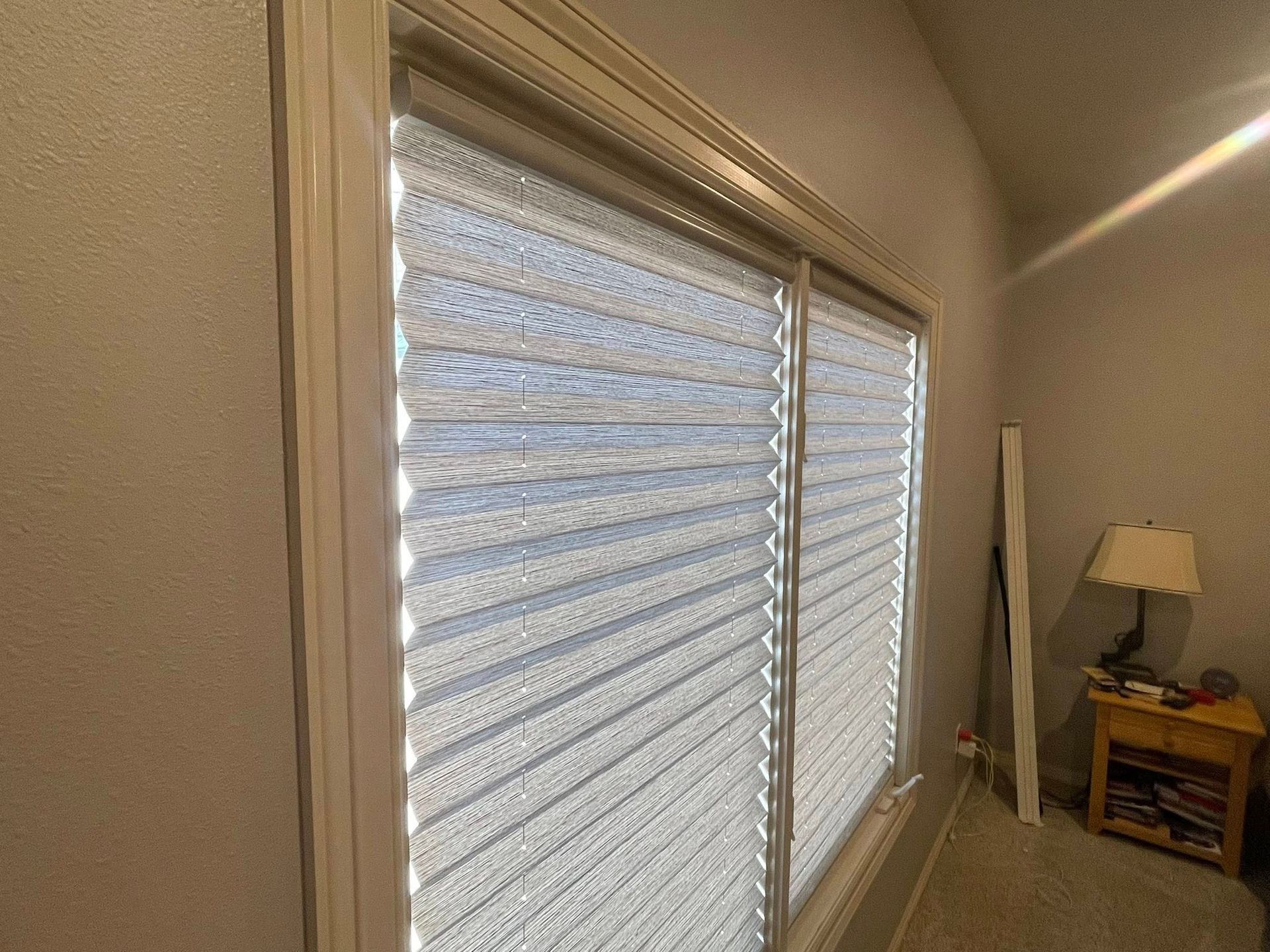 A bright window with horizontal pleated blinds and a wooden side table with a lamp in a corner.