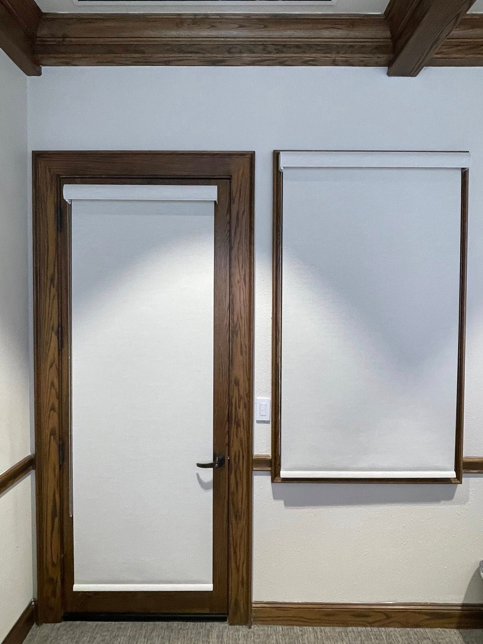 A wooden door with a roller shade next to a window with a matching shade on a light wall under a wooden ceiling beam.
