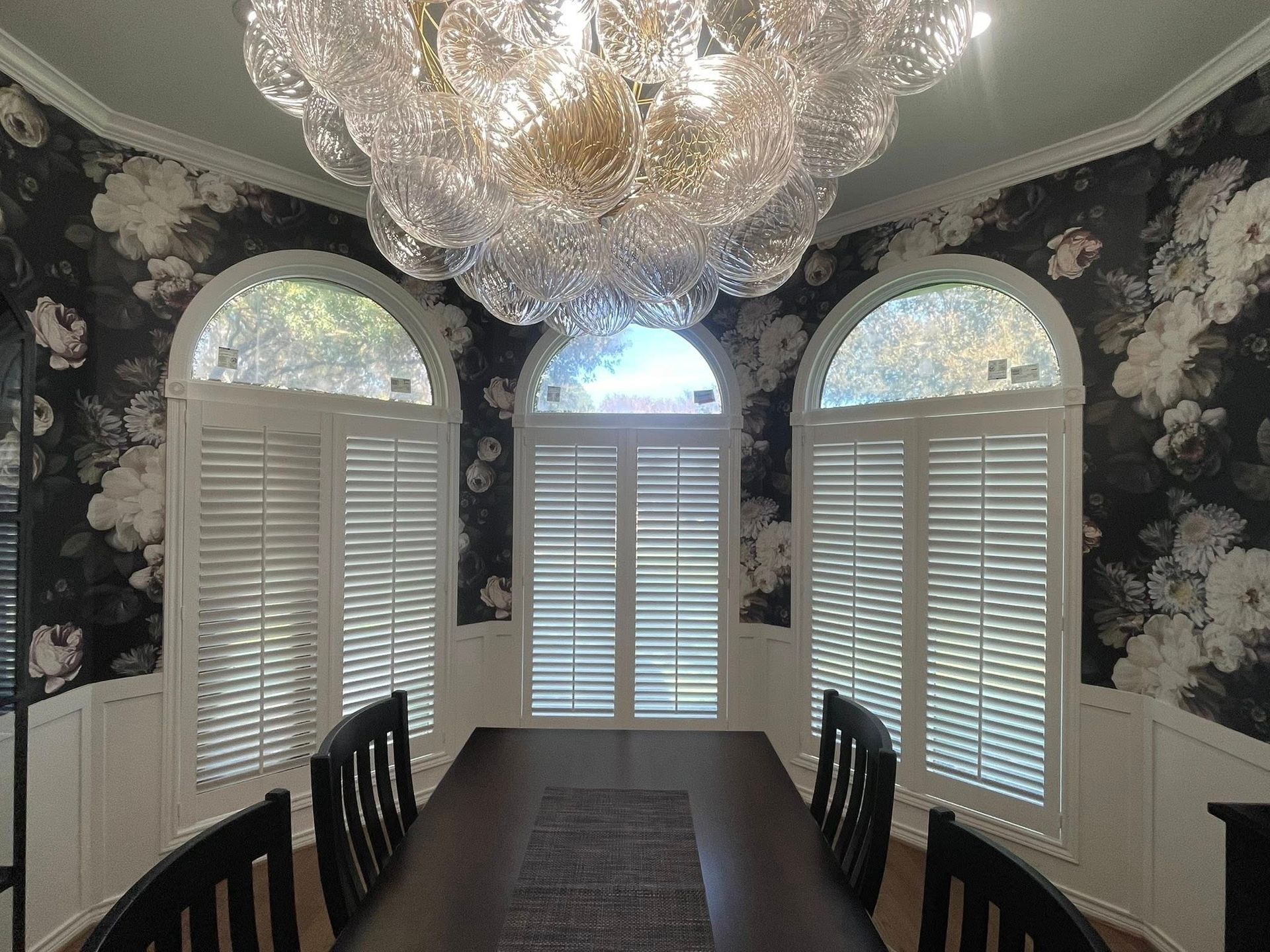 Dining room with a dark floral wallpaper, white shutters on arched windows, and a large, clustered glass chandelier.