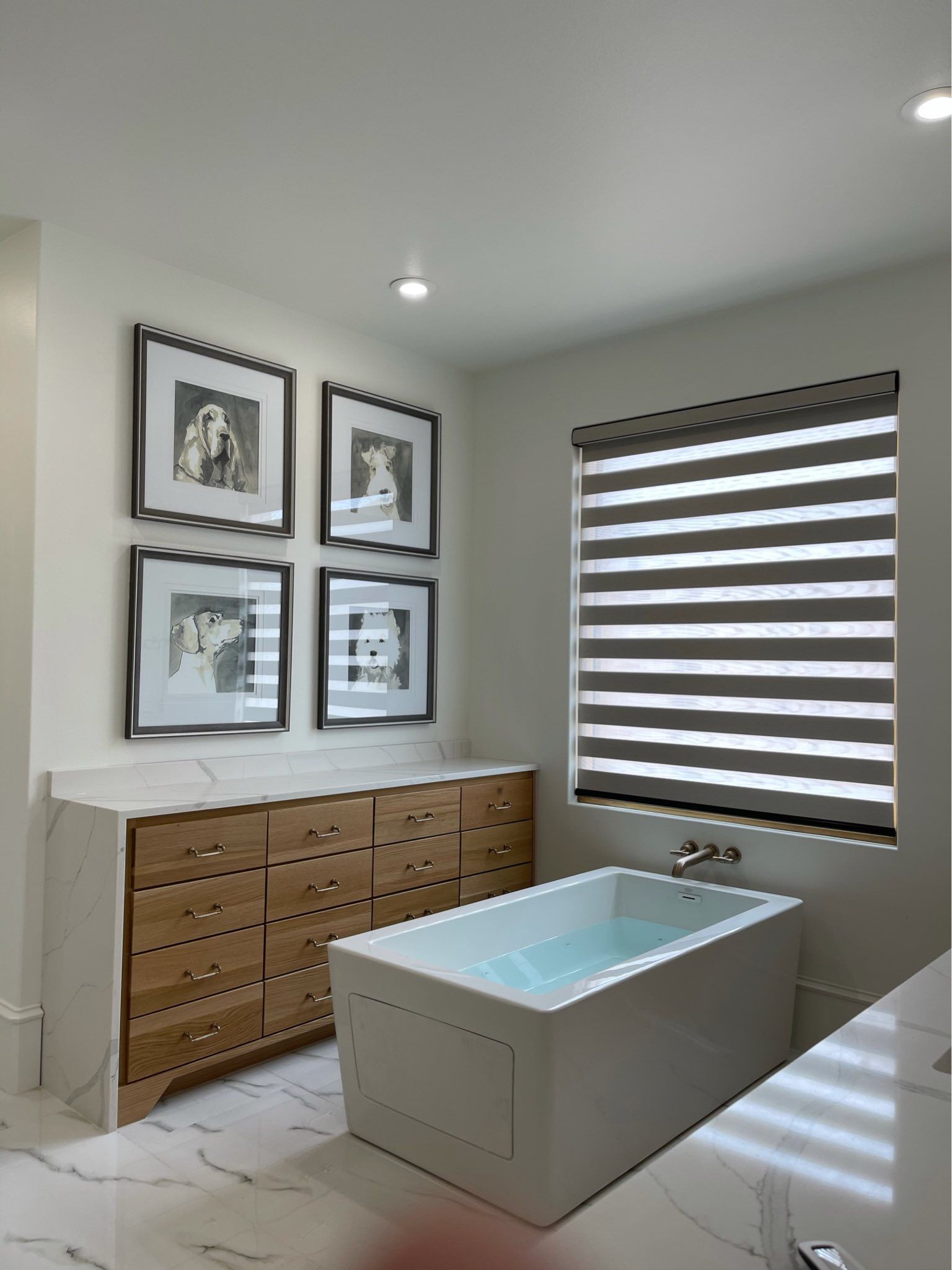 A modern bathroom featuring a white soaking tub, light wood dresser, and four framed black-and-white portraits.