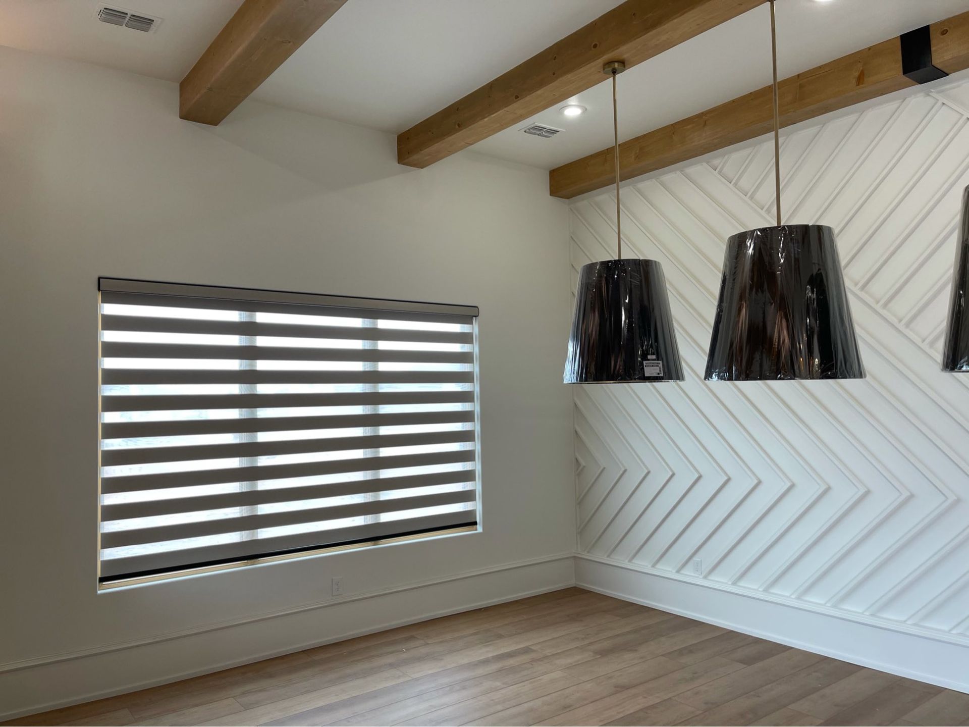 A room featuring a window with striped roller shades, a geometric white accent wall, and wooden beams on the ceiling.