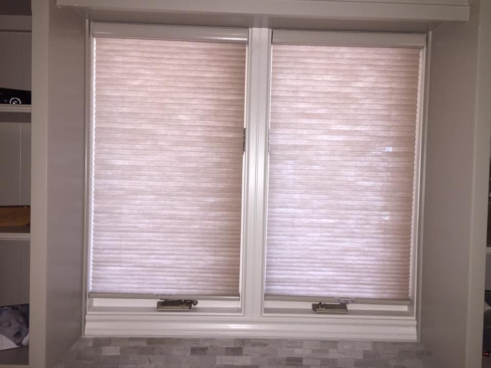 Two light-colored, horizontal pleated shades covering a double window pane above a light gray tiled surface.