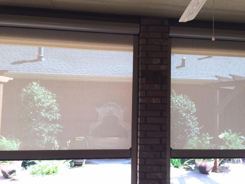 A view through two tan, mesh patio shades showing a brick pillar between them and a garden with a fountain outside.