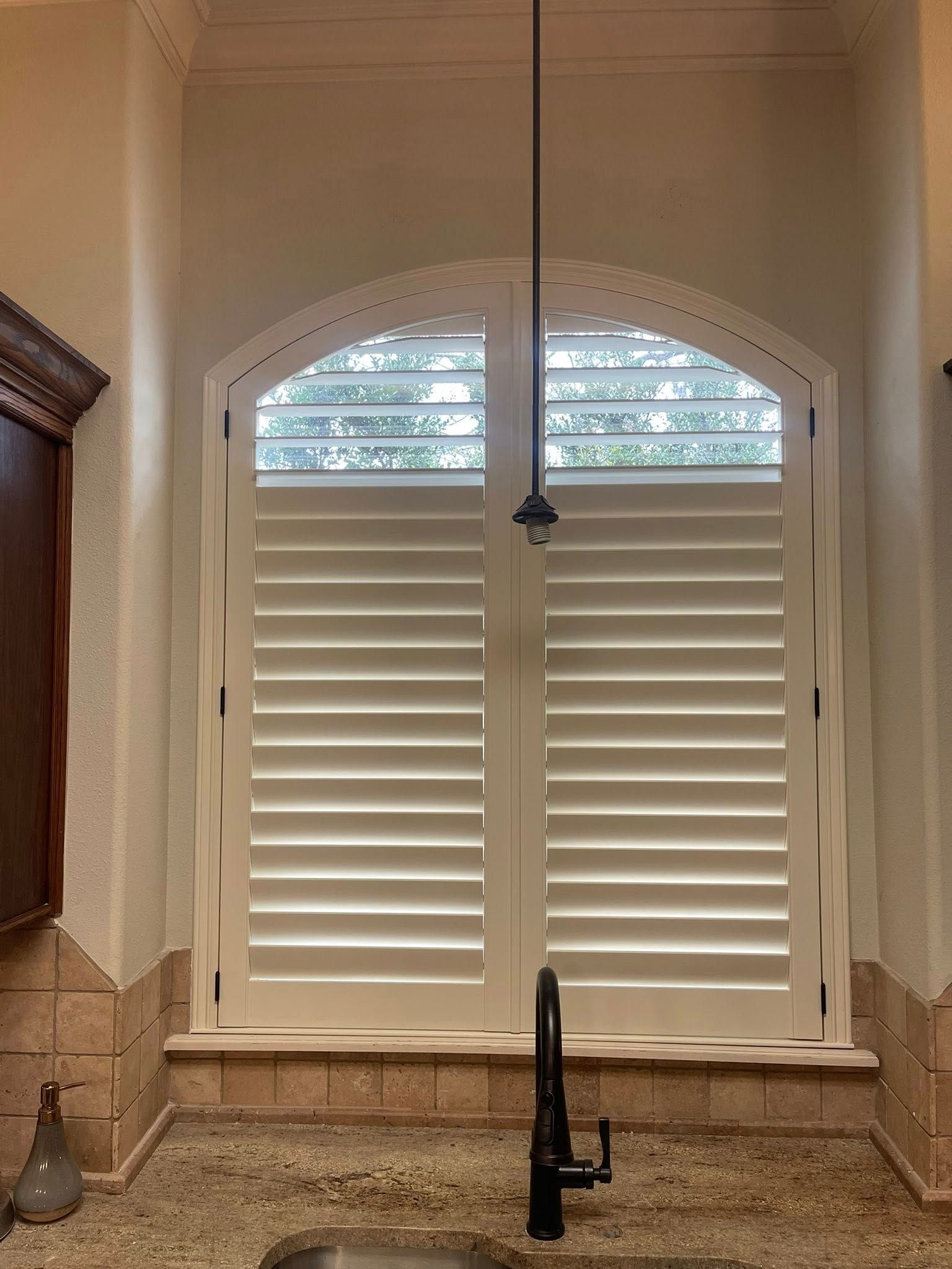 Arched window with white plantation shutters above a kitchen sink, granite countertop, and faucet.