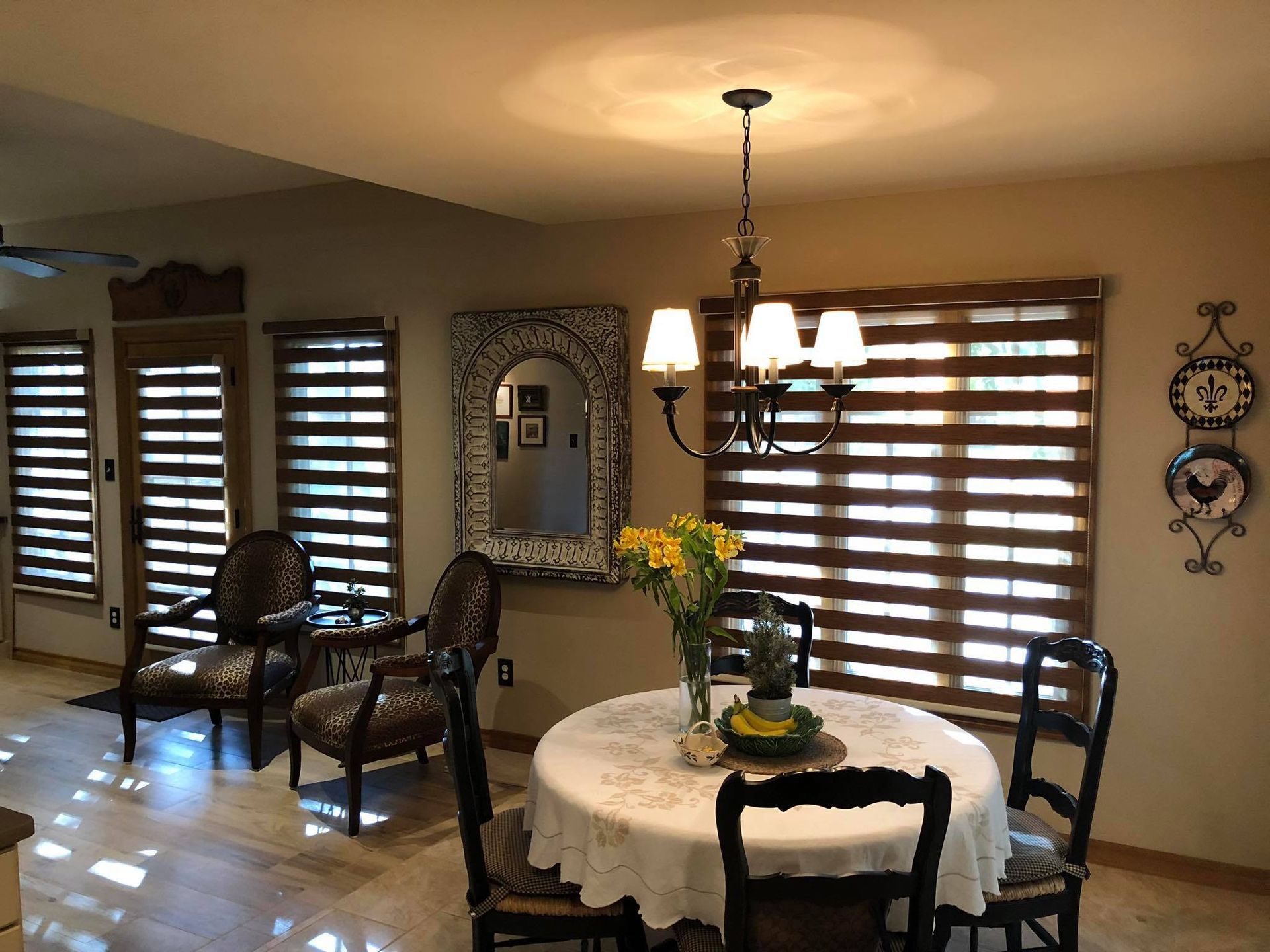 A dining area with a round table, four chairs, a chandelier, a large decorative mirror, and windows with striped blinds.