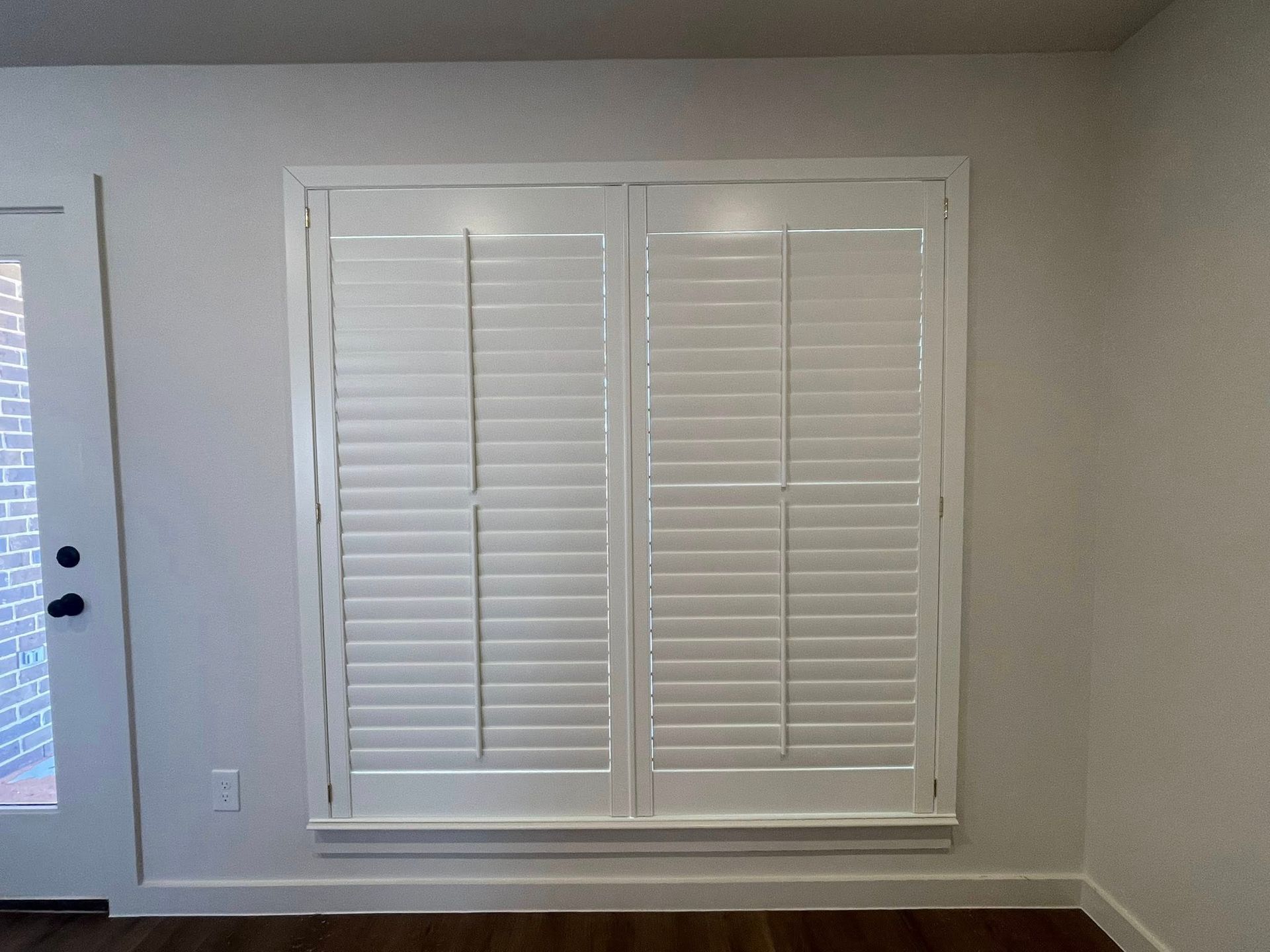 White plantation shutters installed on a large window on a light-colored wall next to a door.