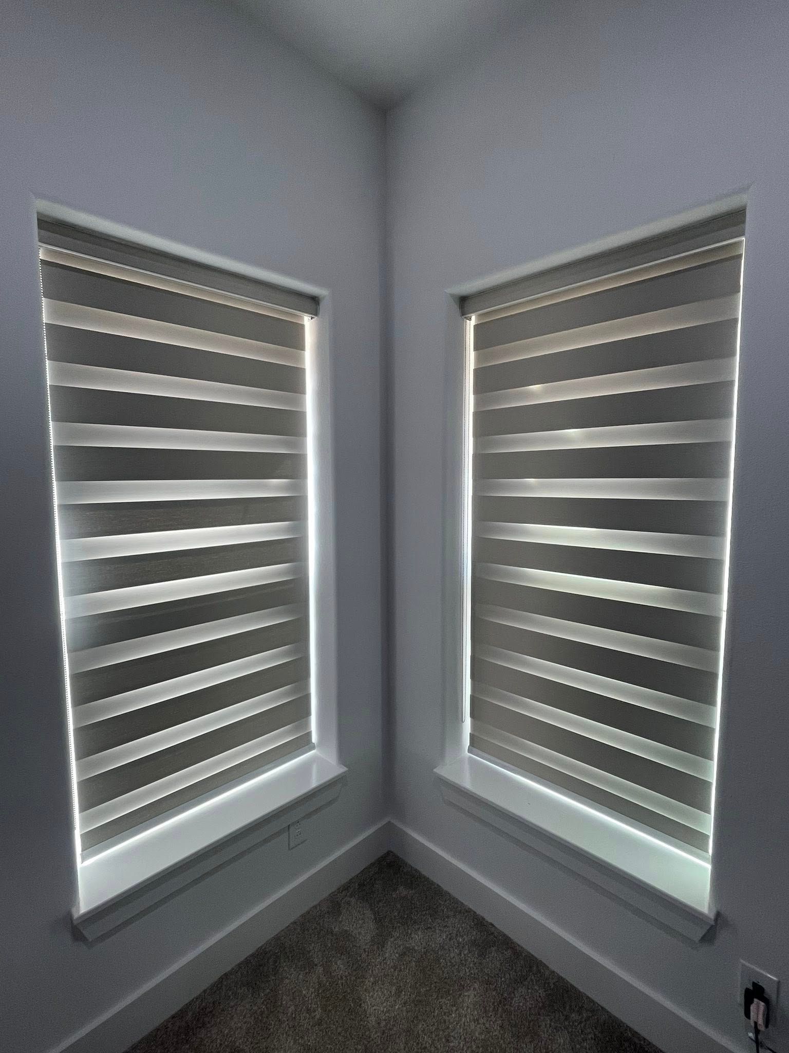 Two windows with grey and white striped zebra blinds in a corner with grey walls and carpeted flooring.