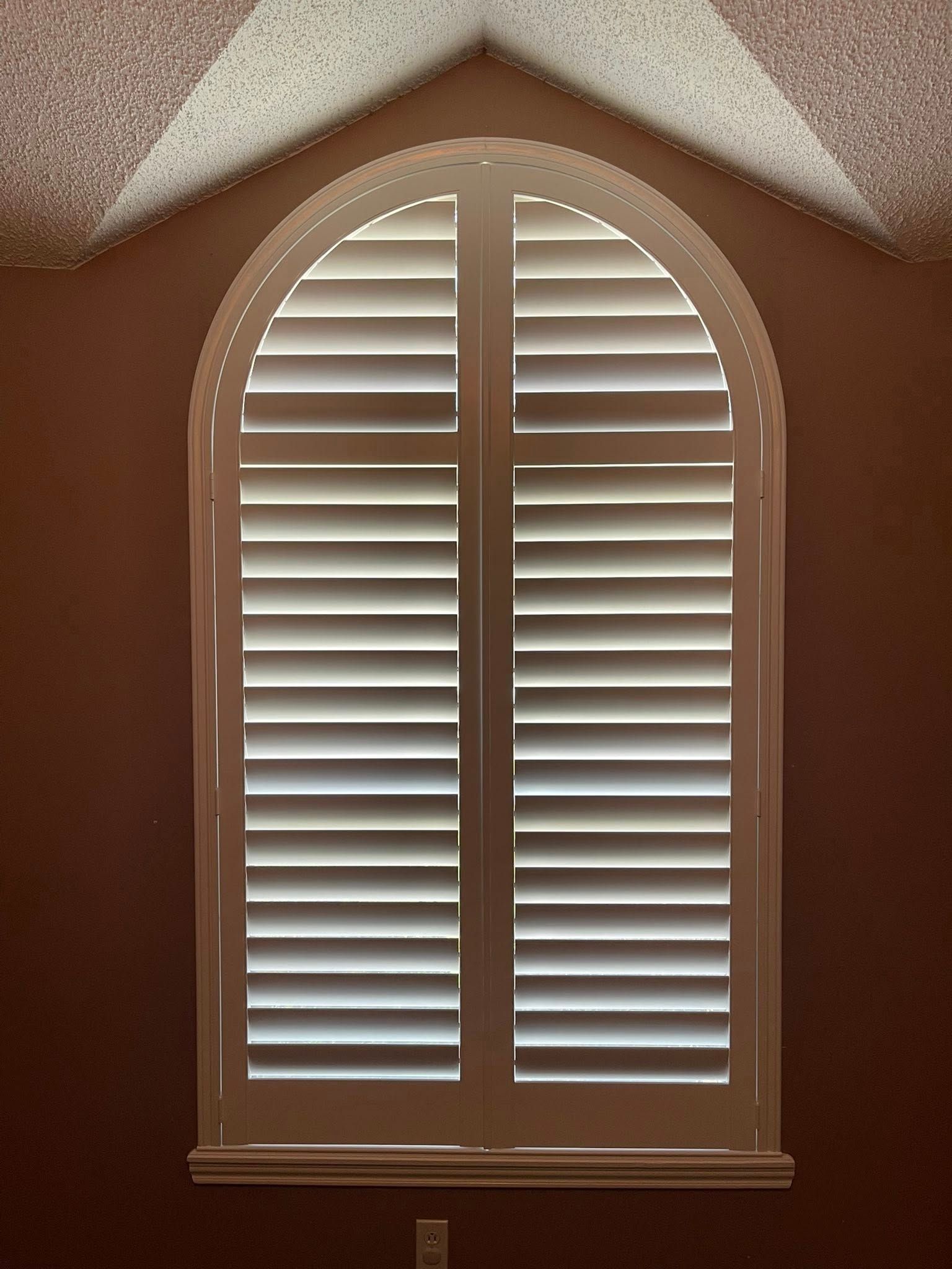 An arched window with white plantation shutters set into a brown wall below a textured, light-colored ceiling peak.