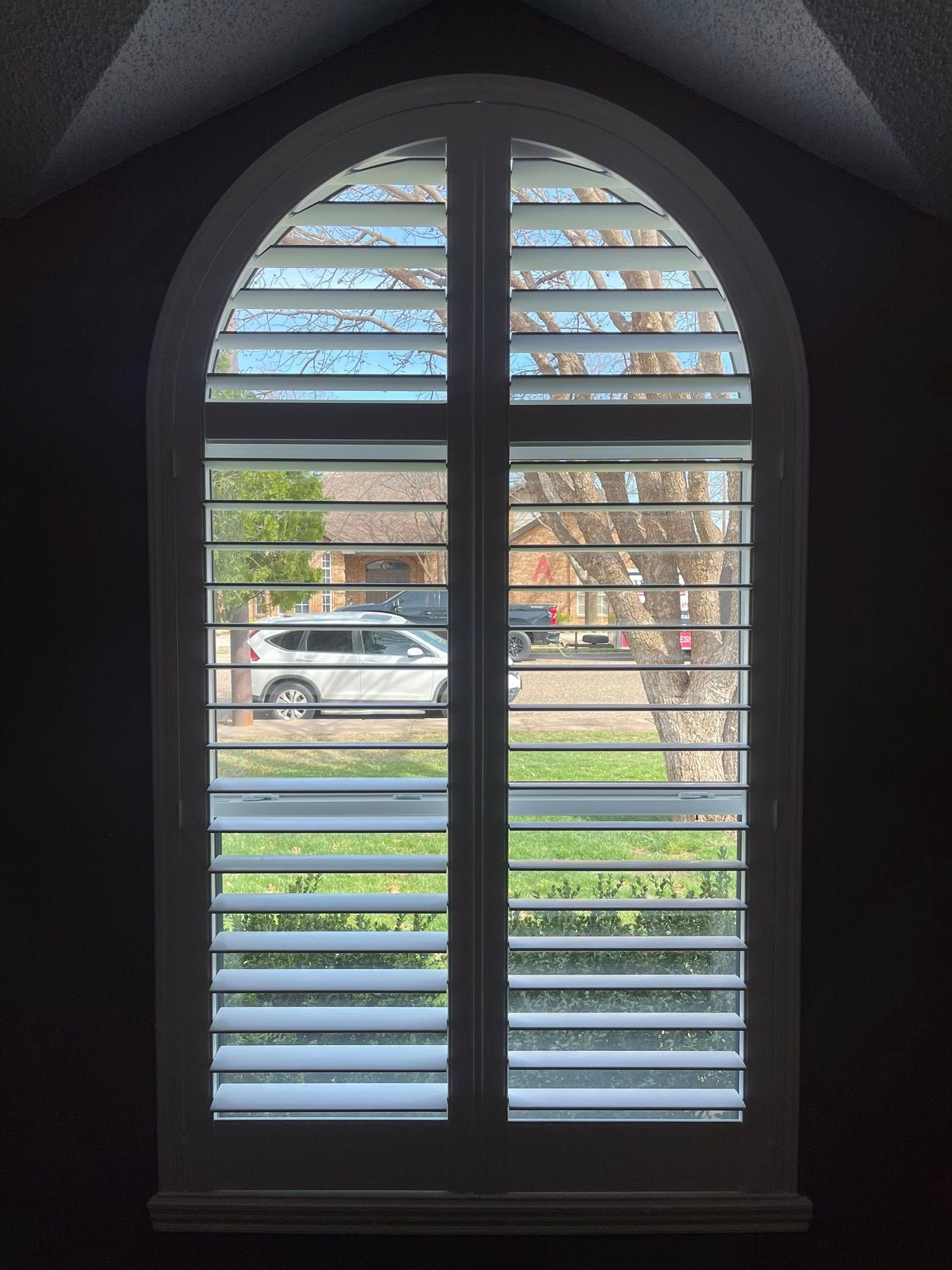 An arched window fitted with white plantation shutters looking out onto a residential street with a car and trees.