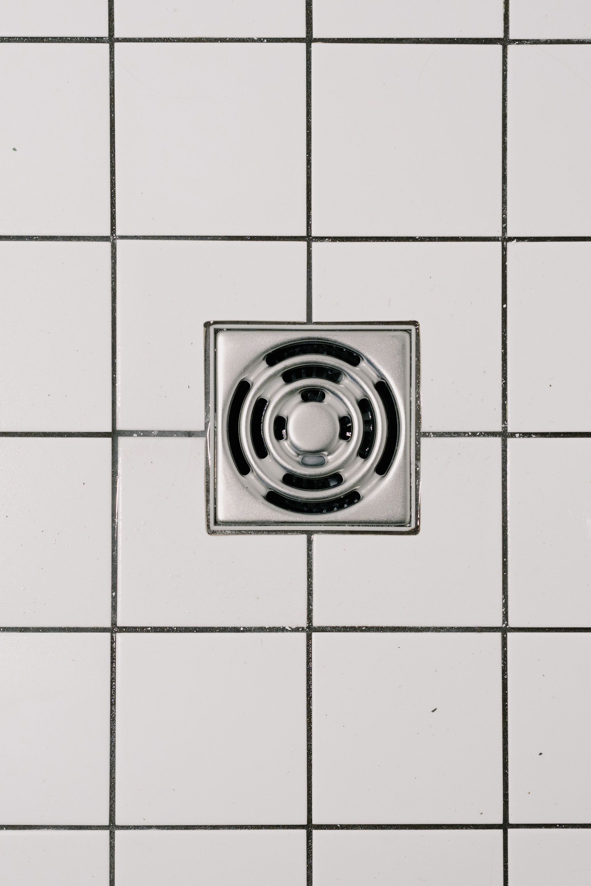 Square metal drain on white tiled wall/floor with a circular grate in the center