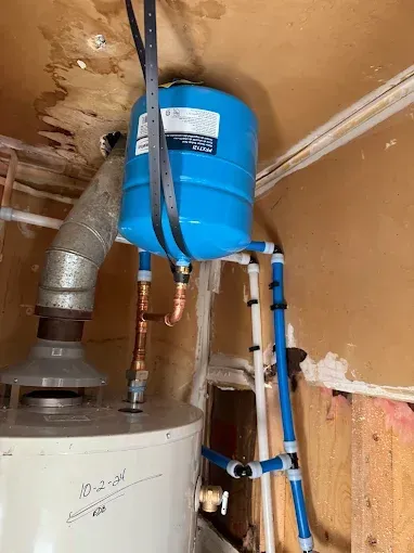 Blue water tank and pipes installed above a water heater in a utility closet, with visible ceiling damage.