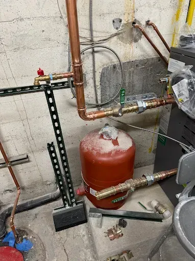 Basement mechanical room with copper pipes, valves, and a red expansion tank beside concrete walls.