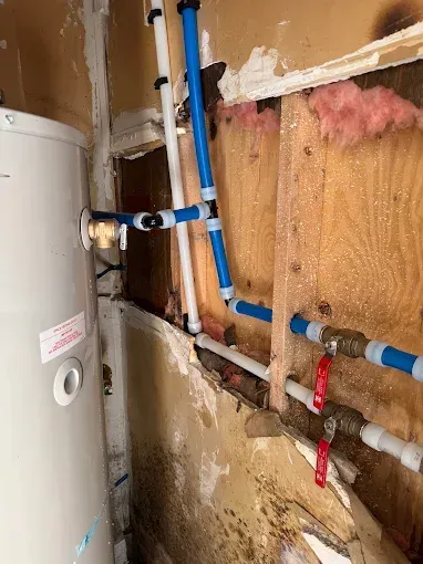 Water heater and exposed blue and copper plumbing inside a damaged wall with pink insulation.