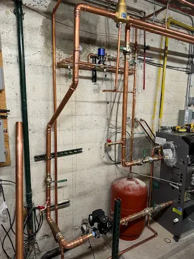 Copper pipes and valves on a basement wall beside a red expansion tank and heating equipment.