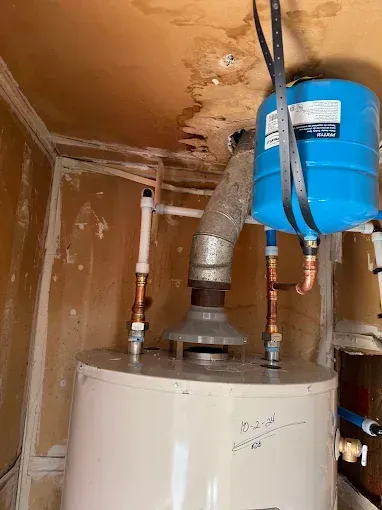 Water heater with a blue expansion tank and vent pipe in a damaged, stained utility closet.