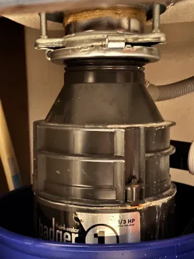 Close-up of a black garbage disposal unit under a sink, with plumbing and metal mounting ring visible.