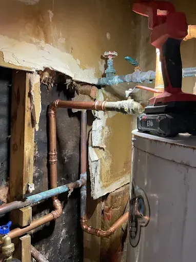 Exposed plumbing and pipes in a damaged wall beside a water heater or boiler unit.