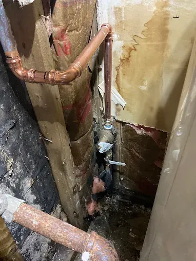 Narrow utility shaft with copper pipes and wet, peeling walls; a worker is partially visible below.